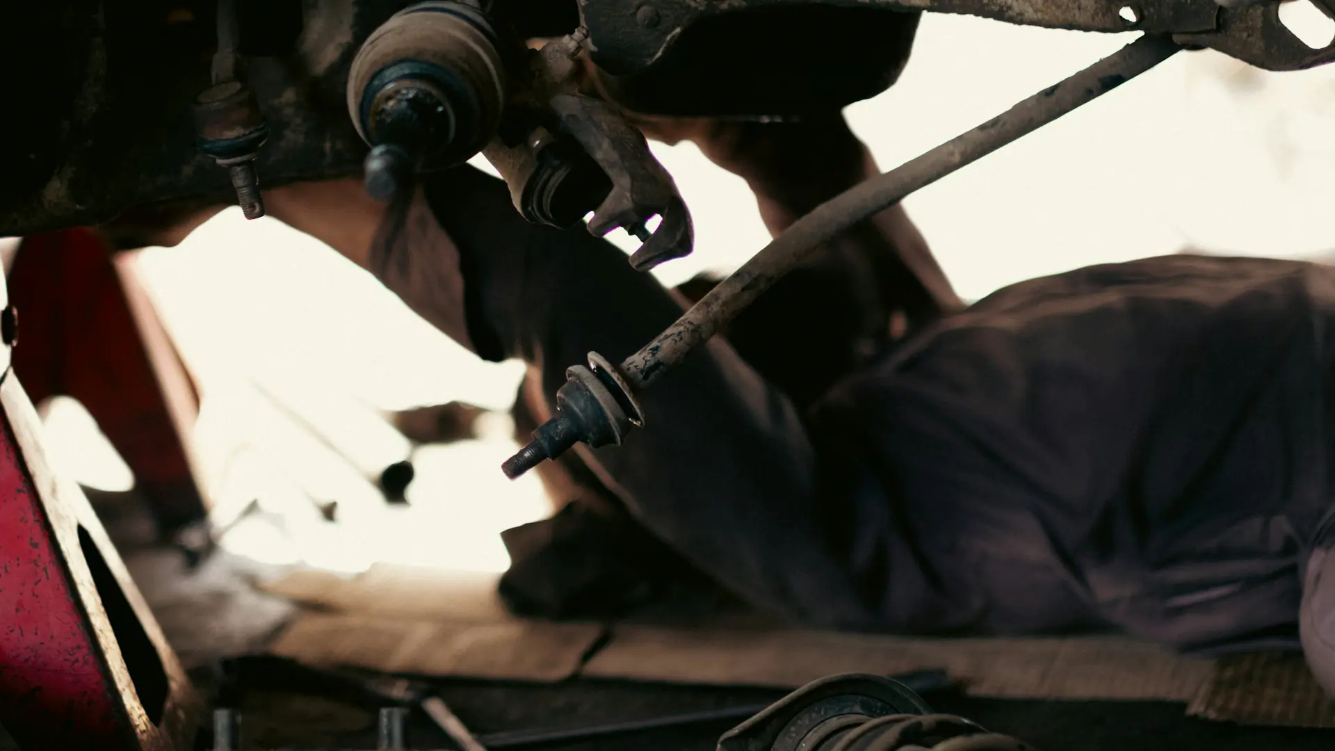 A mechanic lying under a car on cardboard beside a red jack stand.