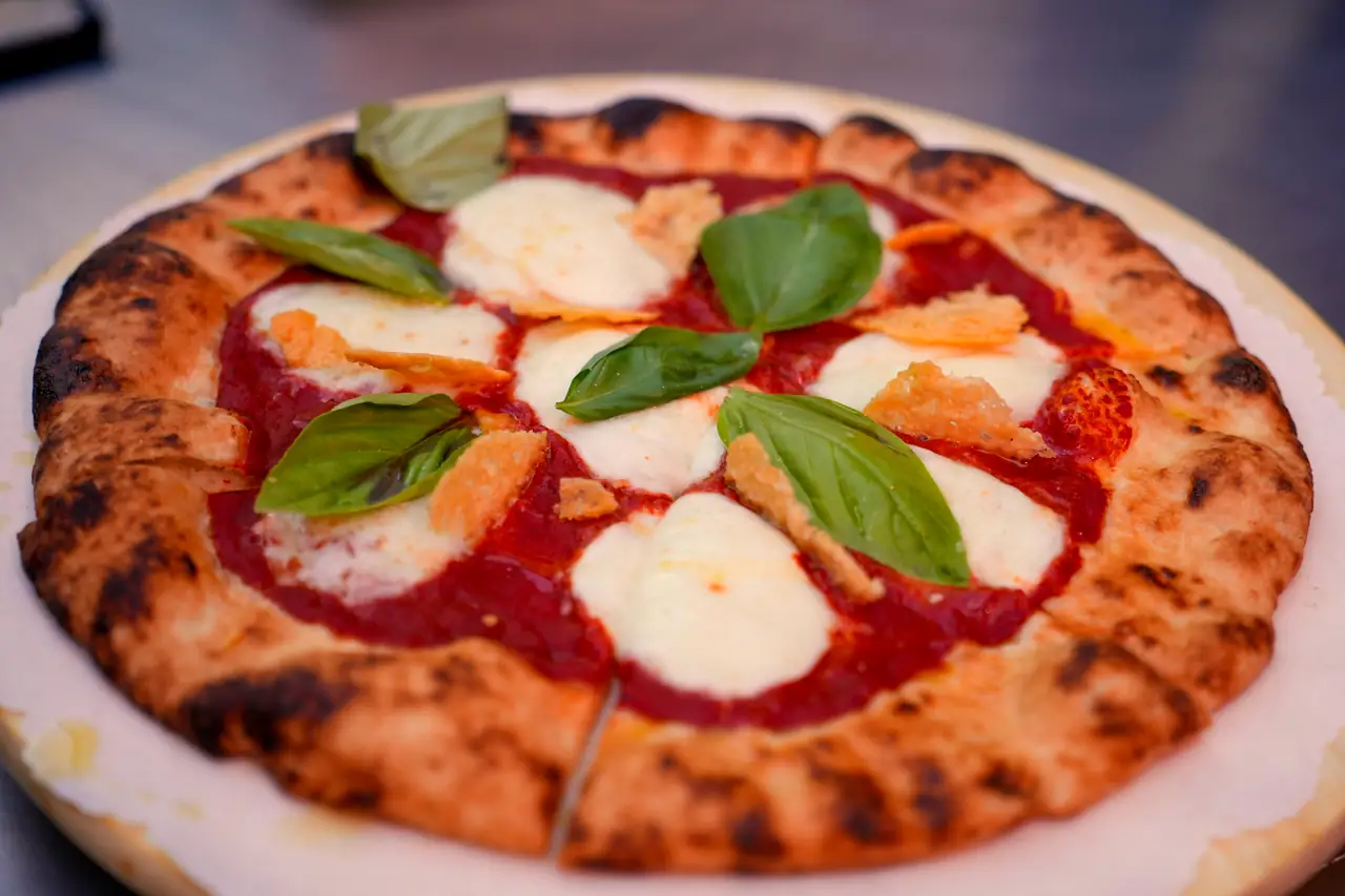 A wood-fired pizza with leopard-charred crust, torn mozzarella and basil, seen from above on the table.