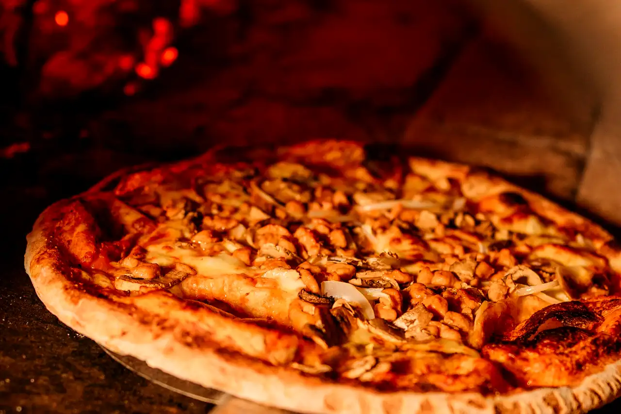 A pie on the peel at the mouth of the wood-fired oven, embers glowing out of focus behind it.