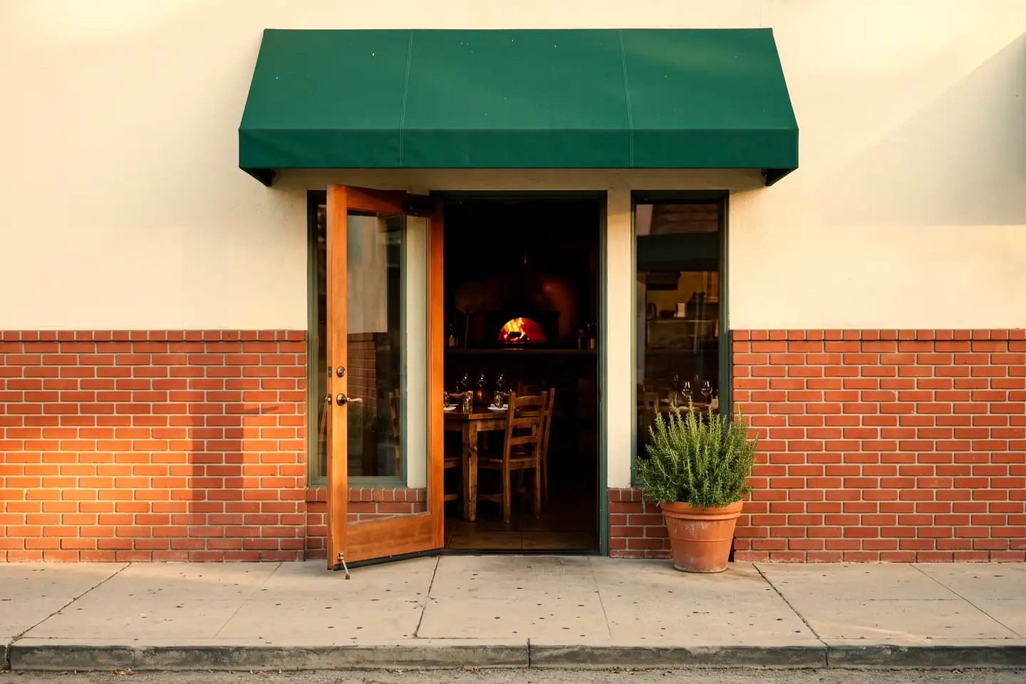 A small storefront with red brick below cream stucco, a dark green awning with no lettering, and a wood-framed glass door open onto a warm dining room with the glow of a wood-fired oven at the back.