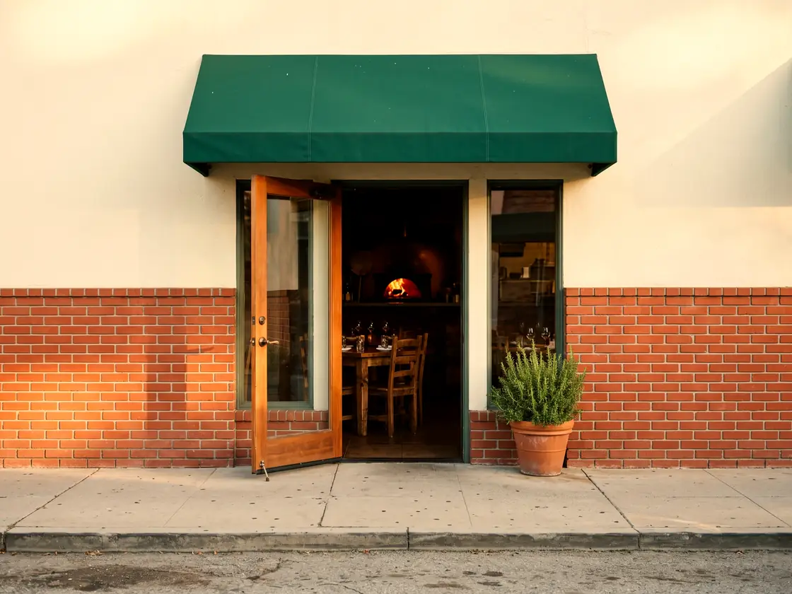 A small storefront with red brick, cream stucco, a green awning and an open door onto a dining room with a wood-fired oven.